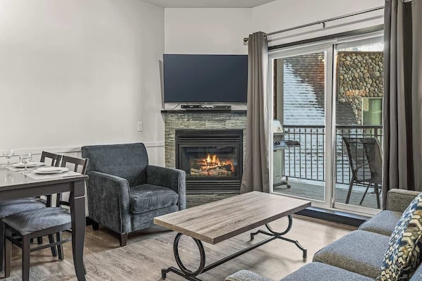 A living room with a fireplace, a flat-screen TV, a dining table with chairs, and a balcony with a view. - Rockies' Embrace- Pool, HotTub, Gym, BBQ, Parking (Canmore)