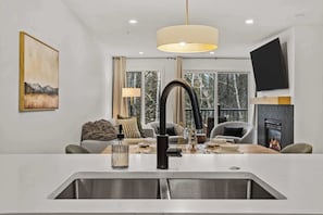 A modern kitchen with a central island, a dining area with a fireplace, and a large window with a view of trees. - Magpie Lookout - Newly Renovated Mountain Escape (Canmore)
