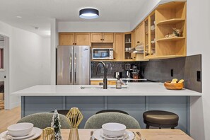 A modern kitchen with a central island, wooden cabinets, stainless steel appliances, and a dining area with a table set for two. - Magpie Lookout - Newly Renovated Mountain Escape (Canmore)