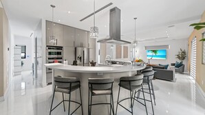 A modern kitchen with a central island, bar stools, stainless steel appliances, and a large TV screen. - Luxury Villa w/ Heated Pool / Jacuzzi / 6BR /Games/ Miami (Miami)