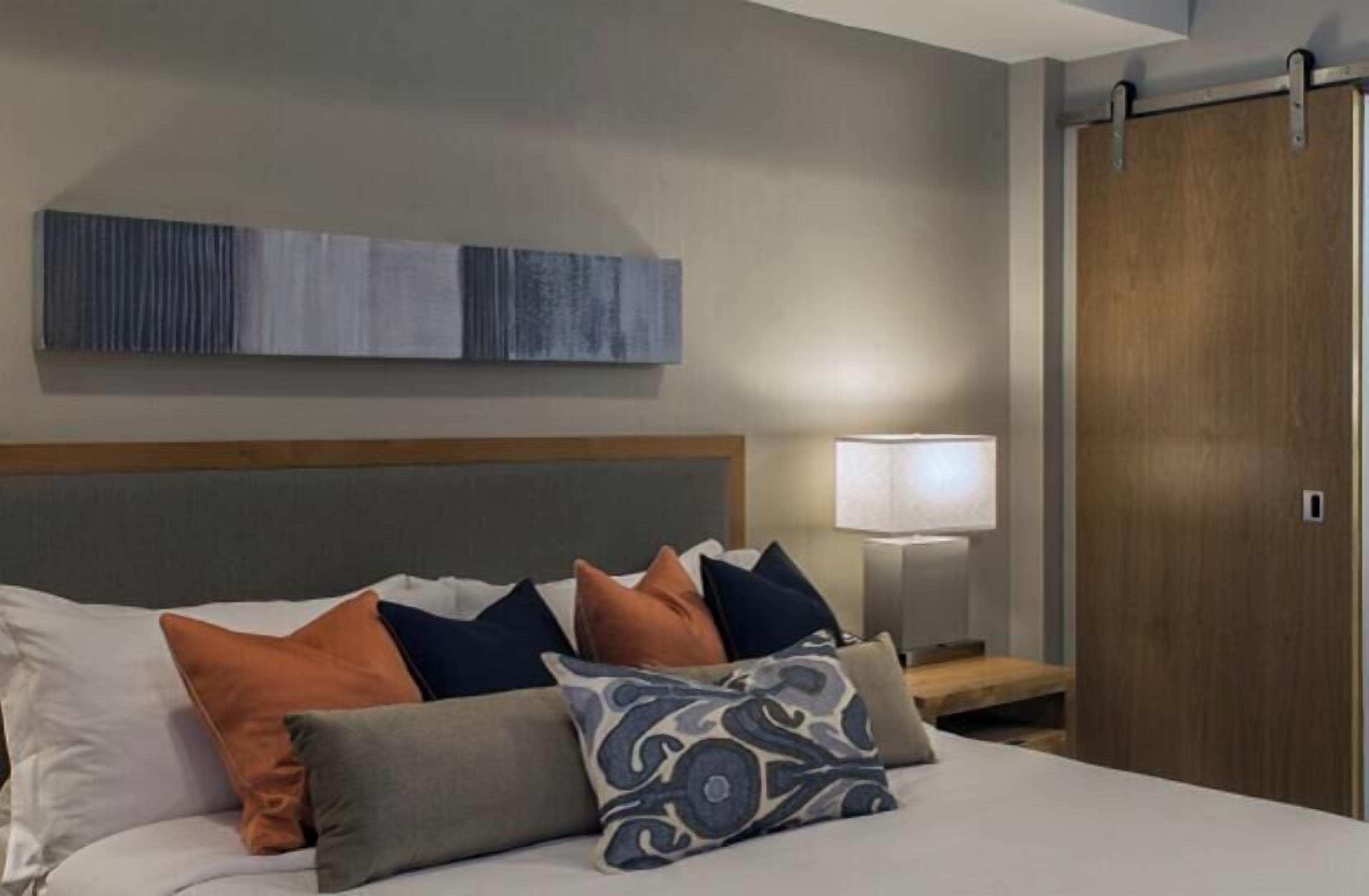 A hotel room with a bed, pillows, a nightstand, a lamp, a wall art piece, and a wooden door.