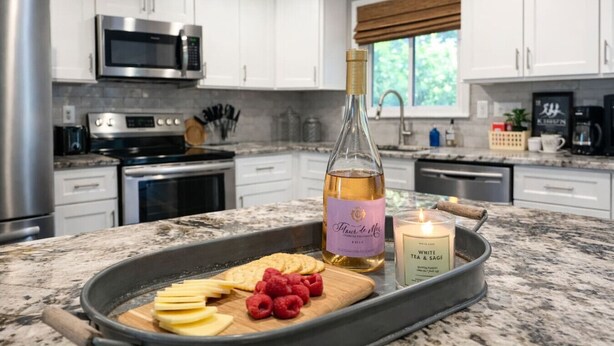 A kitchen with white cabinets, stainless steel appliances, a bottle of wine, a candle, and a tray with cheese and raspberries. - Groups Welcome-Sleeps 20-Pets OK-Fire Pit-BBQ (Charlotte)