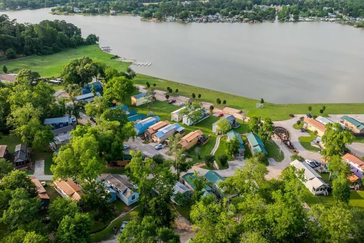 An aerial view of a lakeside campground with RVs and cabins, surrounded by trees and a lake.