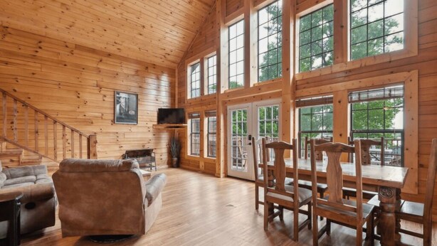 A wooden cabin interior with a staircase, a sofa, a dining table with chairs, and large windows. - Water's Edge RV Resort (Willis)