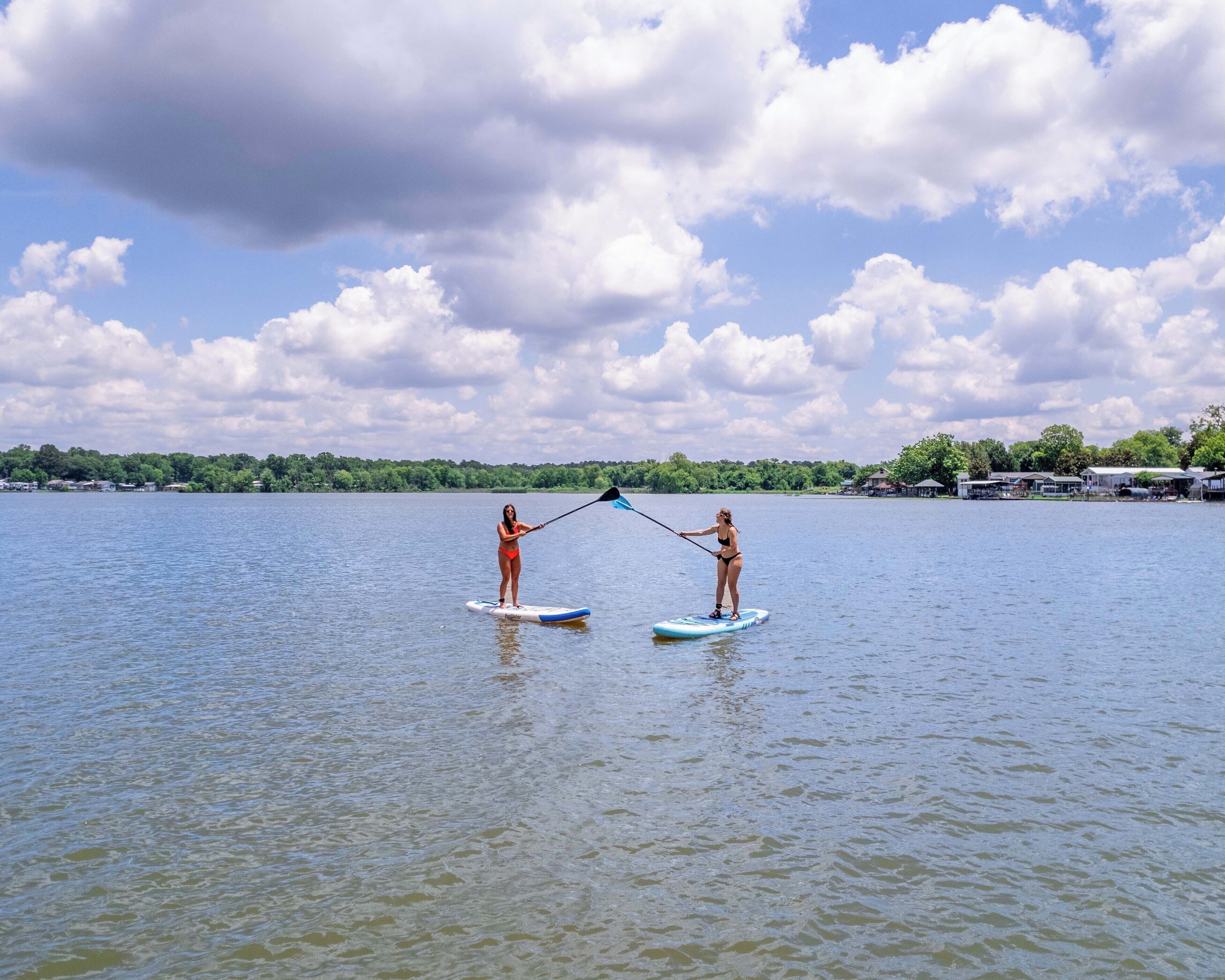 Two people paddleboarding on a calm lake with a clear sky and a tree-lined shore in the background.