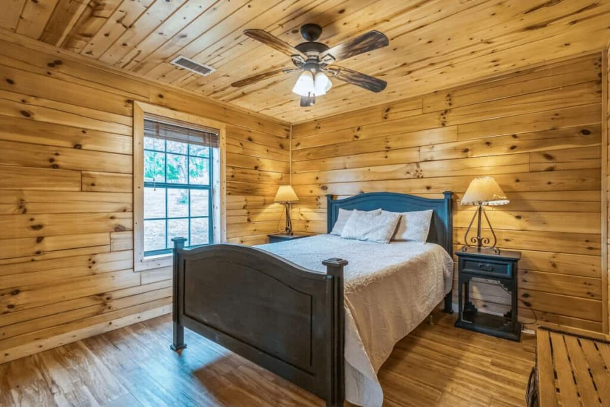 Un dormitorio de cabaña de madera con una cama, dos mesitas de noche, un ventilador de techo y una ventana.