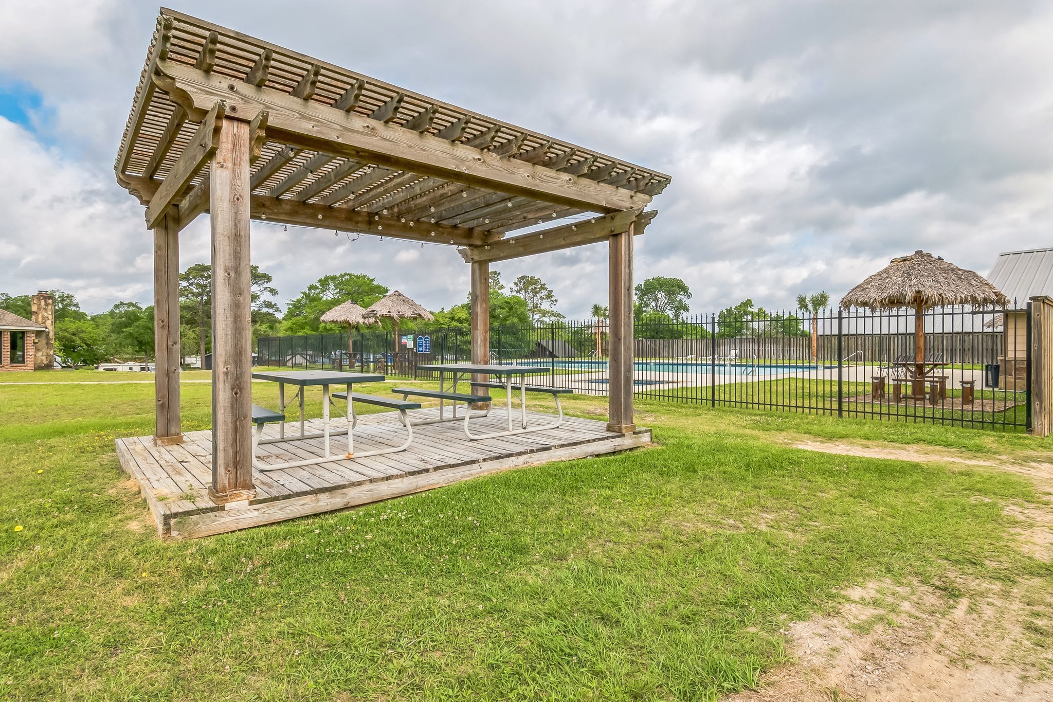 A wooden pergola with benches, a fenced swimming pool, and a thatched-roof structure in the background.