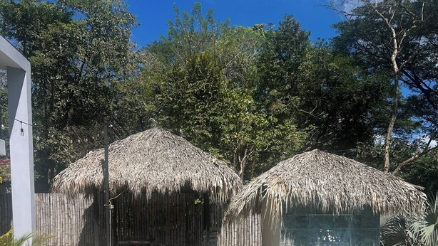 Uma piscina com água cristalina, rodeada por um deck de concreto e duas construções com telhados de palha.