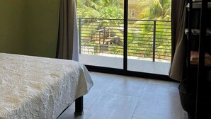 Quarto com cama, ventilador de teto, janela grande com cortinas e vista para o verde lá fora.