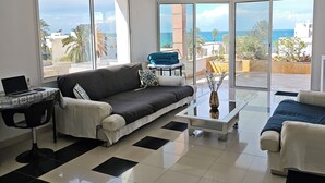 A modern living room with a black and white tiled floor, a grey sofa, a coffee table, and large windows offering a view of the sea.