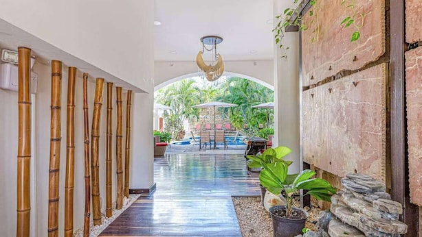 A corridor with a bamboo wall, a stone wall with plants, and a view of an outdoor area with umbrellas and a pool. - Playa Condo, Private Rooftop Terrace & Pool (Playa del Carmen)