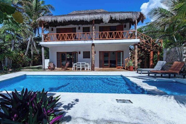 A two-story building with a pool, a thatched roof, and a balcony with wooden railings.