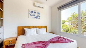 Un dormitorio con cama, mesita de noche, aire acondicionado instalado en la pared, ventana con vistas a la vegetación y un cuadro en la pared.