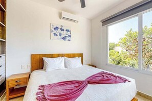 Ein Schlafzimmer mit Bett, Nachttisch, Wand-Klimaanlage, Fenster mit Blick ins Grüne und einem Bild an der Wand.
