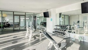 A modern gym with exercise equipment, a mirror, and a television. - Luxury Condo: Rooftop Pool, Jacuzzi & Game Room (Playa del Carmen)