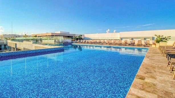 A rooftop swimming pool with lounge chairs, a clear sky, and modern buildings in the background. - Luxury Condo: Rooftop Pool, Jacuzzi & Game Room (Playa del Carmen)