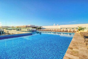 A rooftop swimming pool with lounge chairs, a clear sky, and modern buildings in the background.