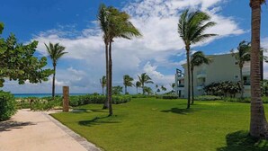 A pathway leading to a building with a view of the ocean, surrounded by palm trees and greenery. - 2BR Beachfront Condo Playa | Pool & Beach (Playa del Carmen)