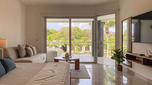 A modern bedroom with a large bed, a sofa, a TV, and a balcony with a view of greenery. - 2BR Beachfront Condo Playa | Pool & Beach (Playa del Carmen)