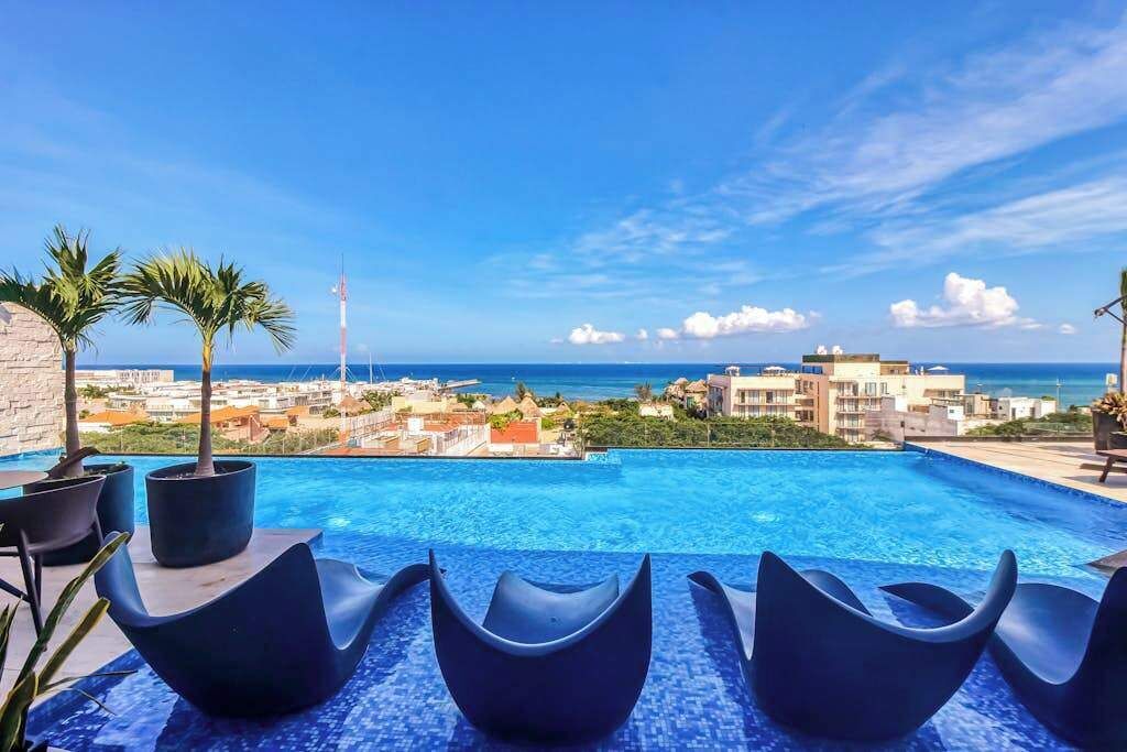 A swimming pool with modern chairs, palm trees, and a view of the ocean and buildings.