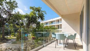 En balkong med ett glasräcke, ett bord med stolar och utsikt över träd och en byggnad.