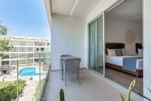 A balcony with a view of a swimming pool, a bed with a wooden headboard, and a dining table with chairs. - Playacar Studio · Resort Pools · Near 5th Ave (Playa del Carmen)