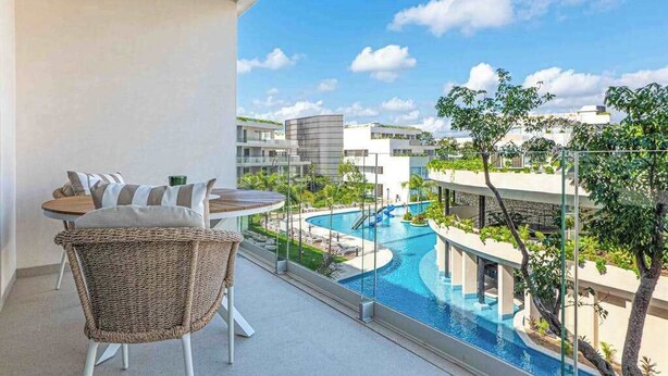 A balcony with a view of a swimming pool, a dining table with chairs, and modern buildings in the background. - Playacar Studio | 4 Pools, Gym, AC & WiFi (Playa del Carmen)