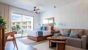 A modern hotel room with a bed, a sofa, a coffee table, a ceiling fan, and a large window. - Playacar Studio | 4 Pools, Gym, AC & WiFi (Playa del Carmen)