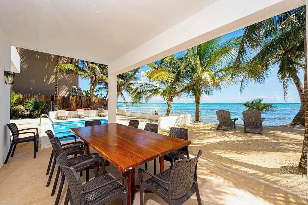 A covered outdoor dining area with a wooden table and chairs, overlooking a beach with palm trees and a view of the ocean.