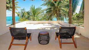 Un área de estar al aire libre techada con dos sillones de mimbre y una mesa redonda, con vista a una playa con palmeras y el océano.