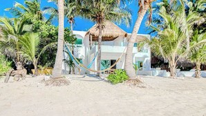 Un hotel frente a la playa con una estructura de techo de paja, palmeras y una hamaca.