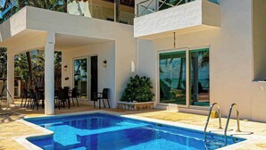 Un área de piscina moderna con una piscina azul cristalina, un espacio de descanso cubierto y vistas al océano a través de amplios ventanales.