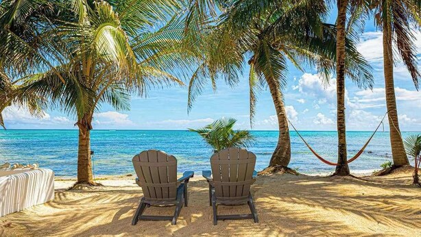 Una playa con dos sillas Adirondack, una hamaca y una mesa blanca, todo bajo palmeras.