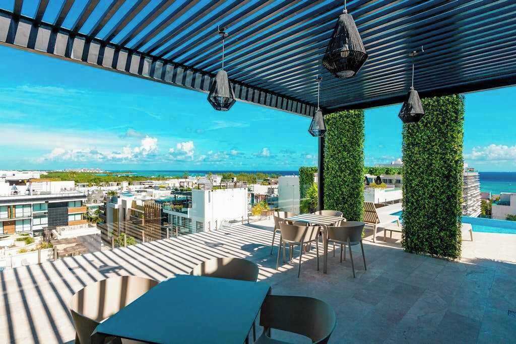 Terraza en la azotea con vistas a la ciudad y al océano, que incluye una mesa y sillas, luces colgantes y jardines verticales verdes.