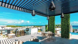 A rooftop terrace with a view of the city and ocean, featuring a table and chairs, hanging lights, and green vertical gardens. - Studio | Rooftop Pool & Gym, Steps to Beach (Playa del Carmen)