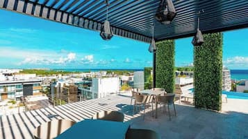 Terraza en la azotea con vistas a la ciudad y al océano, que incluye una mesa y sillas, luces colgantes y jardines verticales verdes.