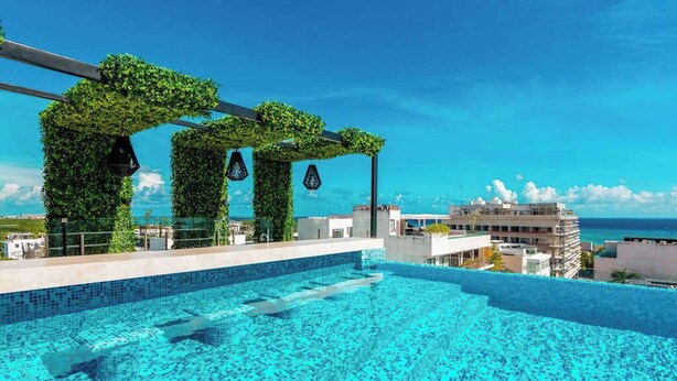 A rooftop infinity pool with a clear view of the ocean, surrounded by greenery and modern architecture. - Studio | Rooftop Pool & Gym, Steps to Beach (Playa del Carmen)