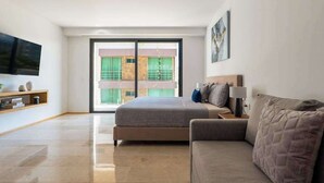 A modern bedroom with a bed, a sofa, a TV, and a balcony with a view of buildings. - Studio | Rooftop Pool & Gym, Steps to Beach (Playa del Carmen)