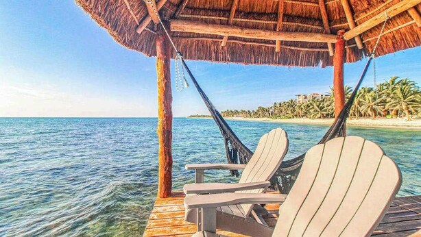 A wooden deck with two Adirondack chairs under a thatched roof, overlooking a clear blue sea and a beach with palm trees. - Beachfront 7BR Villa with Private Pool and Sauna (Tulum)