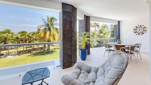 A balcony with a view of palm trees, a grey cushioned chair, a round table with chairs, and a blue potted plant. - Mareazul Beachfront 2BR | Pool & Sea Views (Playa del Carmen)