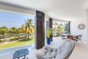 A balcony with a view of palm trees, a grey cushioned chair, a round table with chairs, and a blue potted plant. - Mareazul Beachfront 2BR | Pool & Sea Views (Playa del Carmen)