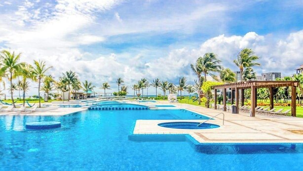 A large outdoor swimming pool with multiple sections, surrounded by palm trees and a covered seating area. - Mareazul Beachfront 2BR | Pool & Sea Views (Playa del Carmen)