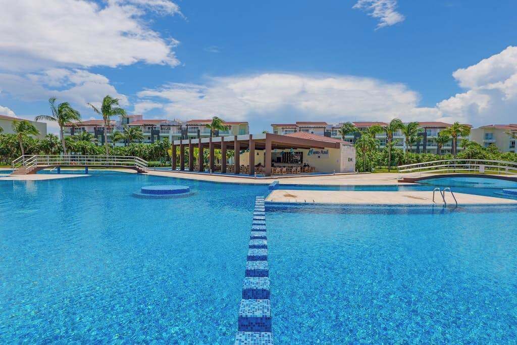 A large outdoor swimming pool with clear blue water, surrounded by palm trees and hotel buildings.