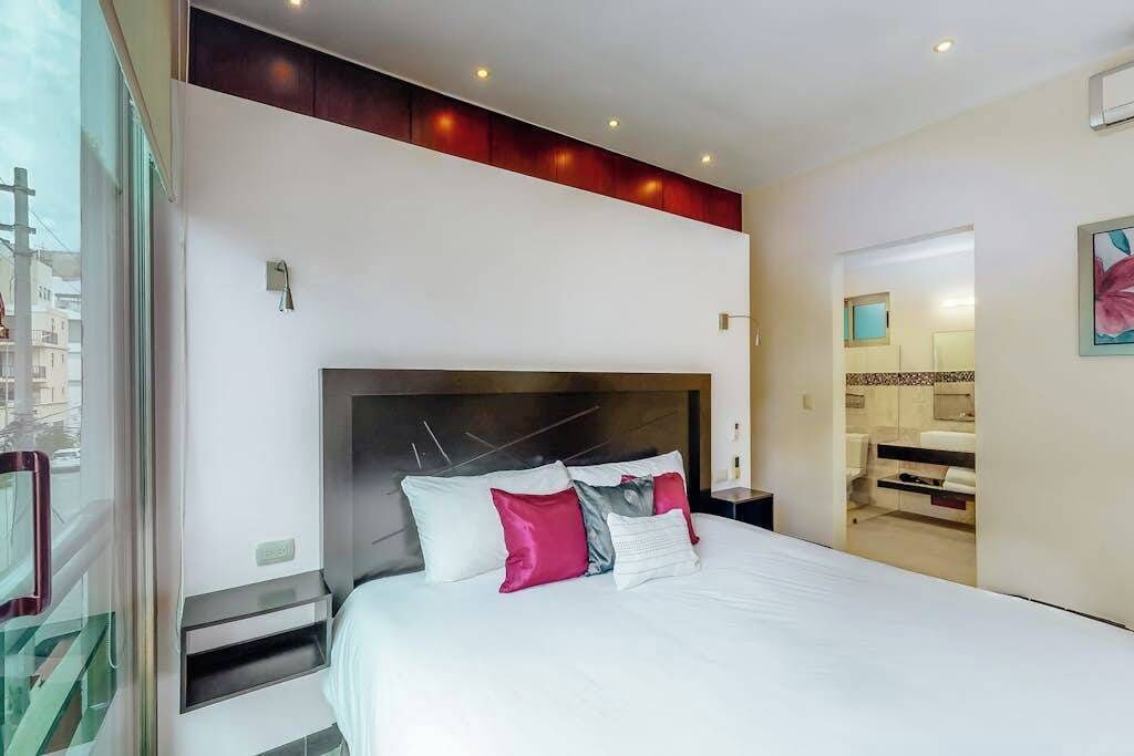 A modern hotel room with a large bed, a headboard with light fixtures, a bedside table, and a view of the city through a window.
