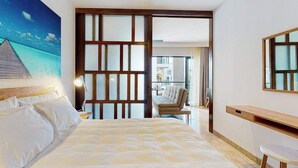 Una camera d'albergo con un letto, una scrivania, una sedia, una grande finestra e un quadro raffigurante una spiaggia.