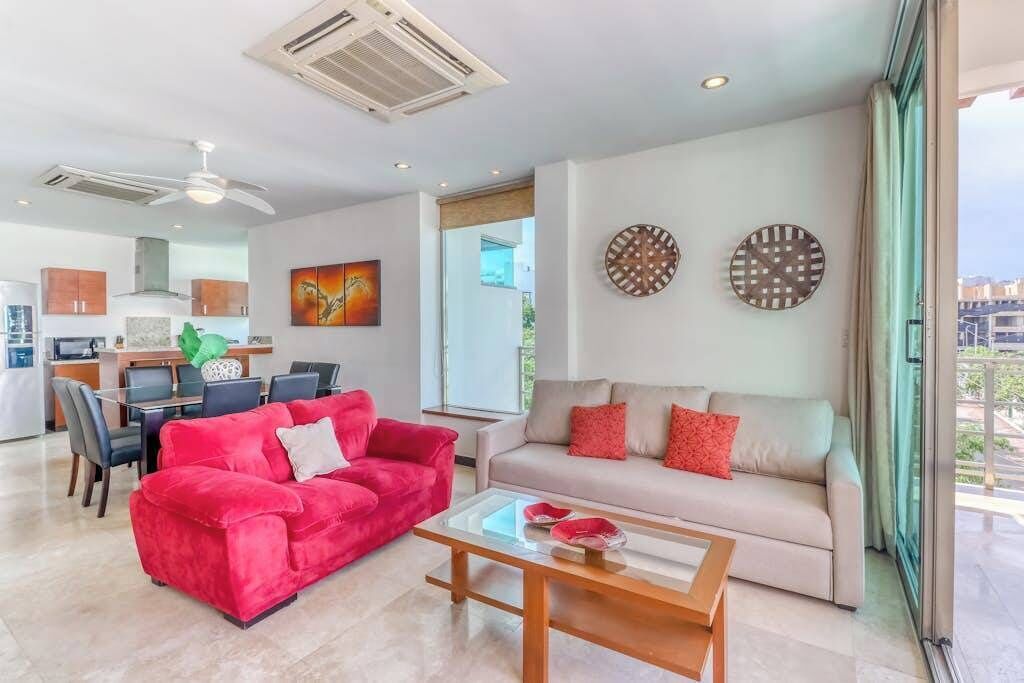 A modern living room with a red sofa, a beige sofa, and a coffee table. There is a dining area with chairs and a table, and a kitchen in the background.