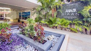 Ein moderner Hotel Eingang mit einem angelegten Garten, einem Schild mit dem Schriftzug 'SIAAN' und Blick ins Innere des Gebäudes.