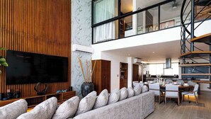 Ein modernes Wohnzimmer mit großem Flachbildfernseher, einer Couch mit vielen Kissen, einer Wand mit Holzdekor und einem Essbereich mit Tisch und Stühlen.