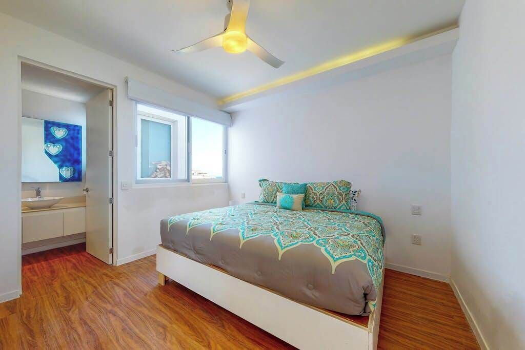 Een slaapkamer met een bed, een plafondventilator, een raam en een badkamer met een spiegel.