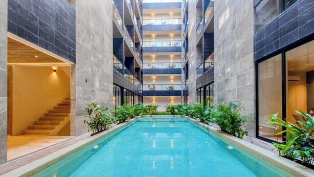 A modern indoor swimming pool with a central staircase, surrounded by plants and with a high ceiling. - Modern 1BR Condo | Rooftop Pool & Gym, Near Beach (Playa del Carmen)